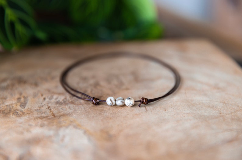 May include: A minimalist bracelet featuring three speckled, off-white beads strung on a thin, dark brown leather cord. The cord is knotted at each end. The bracelet is displayed on a wooden surface.