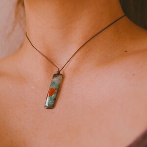 May include: A rectangular pendant necklace with a teal, red, and green design. The pendant hangs from a thin, dark brown cord. The necklace is worn around the neck.