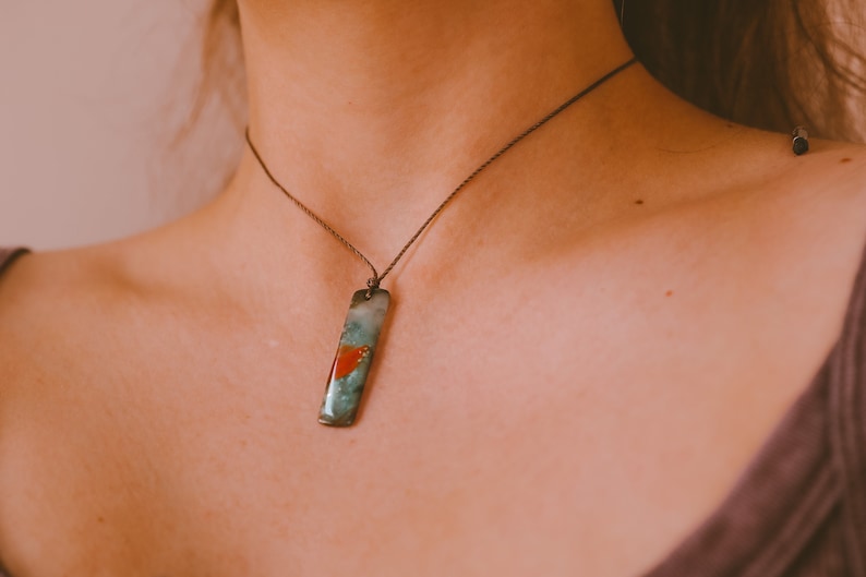 May include: A rectangular pendant necklace with a teal, red, and green design. The pendant hangs from a thin, dark brown cord. The necklace is worn around the neck.