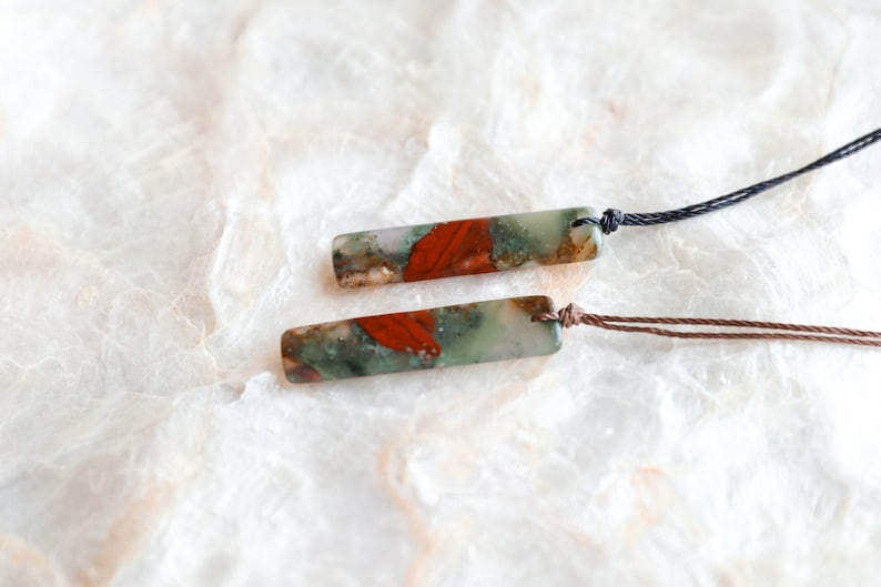 May include: Two rectangular pendant necklaces. Each pendant features a mix of green, red, and brown tones. One necklace has a black cord, the other a brown cord. The pendants are displayed on a textured, off-white surface.