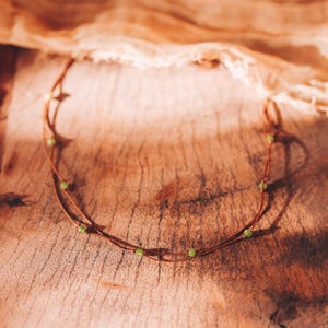 May include: A delicate necklace with a thin, brown cord and small, round, light green beads. The necklace is presented on a textured wooden surface, creating a natural aesthetic. The beads are evenly spaced along the cord, giving a minimalist look.