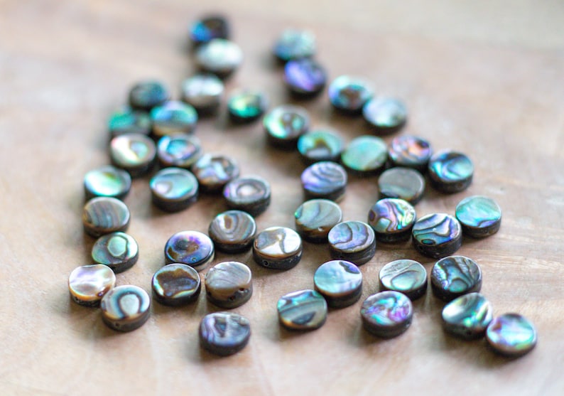May include: A collection of iridescent abalone shell beads. The beads are round and flat, displaying a range of colors including blue, green, purple, and brown. These beads are ideal for jewelry making and crafting projects.