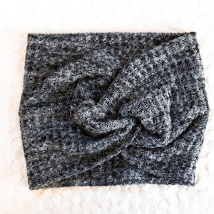 May include: A grey and white knitted headband with a twisted knot detail. The headband is made from a textured fabric and is shown on a white surface. This accessory is designed to keep hair away from the face.