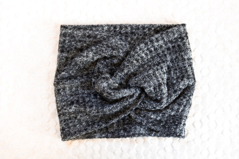 May include: A grey and white knitted headband with a twisted knot detail. The headband is made from a textured fabric and is shown on a white surface. This accessory is designed to keep hair away from the face.