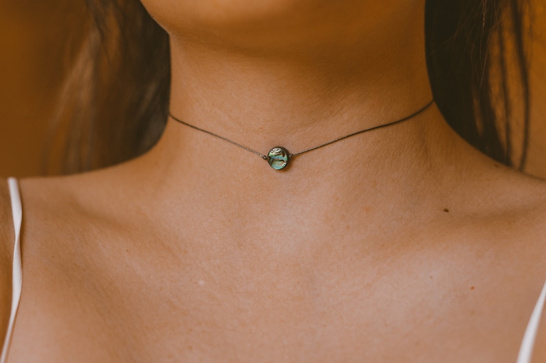 May include: A delicate choker necklace featuring a small, round pendant with iridescent green and blue hues. The pendant is suspended from a thin, dark-colored chain. The necklace is worn around the neck.