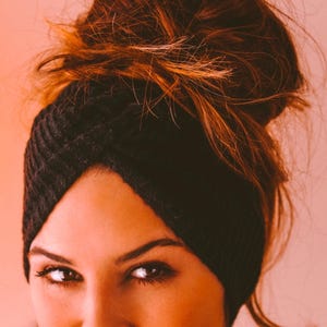 Peut inclure: Gros plan sur une femme portant un turban noir en tricot. Le turban est enroulé autour de la tête, couvrant les oreilles et le front. Les cheveux bruns de la femme sont relevés en un chignon en désordre. Le fond est orange clair.