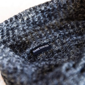 May include: Close-up of a grey and black knitted beanie. The beanie has a textured pattern and a small black tag that reads "SAMESUN CO" in white letters. The fabric appears soft and warm.