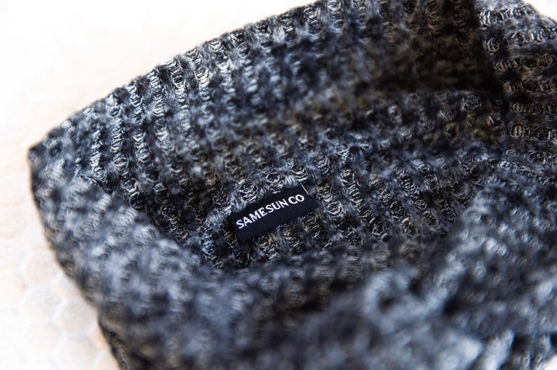 May include: Close-up of a grey and black knitted beanie. The beanie has a textured pattern and a small black tag that reads "SAMESUN CO" in white letters. The fabric appears soft and warm.