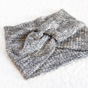 May include: A grey and white knitted headband with a twisted knot detail. The headband is made of a textured fabric and is shown on a white surface. This accessory is suitable for various hairstyles.