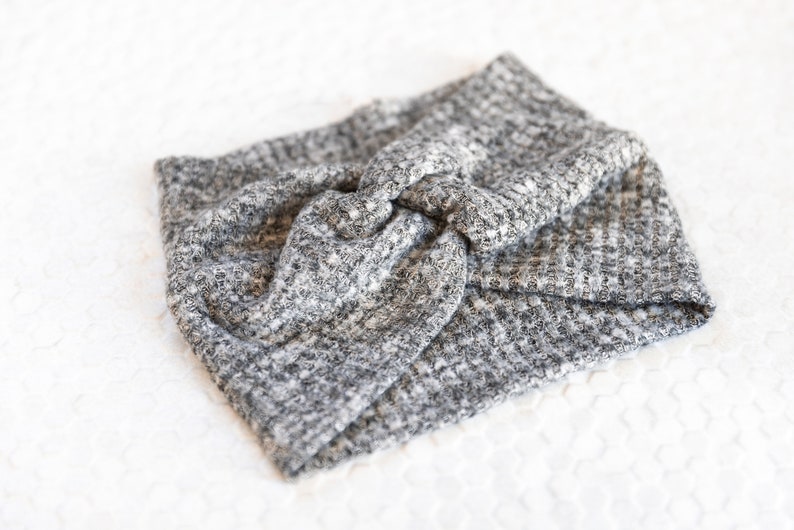 May include: A grey and white knitted headband with a twisted knot detail. The headband is made of a textured fabric and is shown on a white surface. This accessory is suitable for various hairstyles.