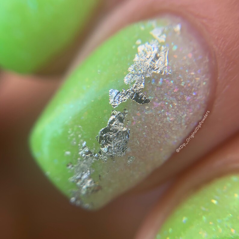 May include: Close-up of a green nail with silver flakes and white glitter on top. The nail is surrounded by a light green background. The text "@KDIP_OriginalDipPowders" is visible in the lower right corner of the image.