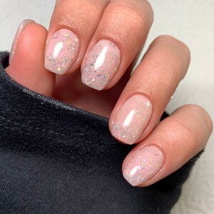 K&ouml;nnte beinhalten: Nahaufnahme einer Hand mit f&uuml;nf N&auml;geln, die mit einem hellrosa Nagellack mit schimmerndem Glitzer lackiert sind. Die N&auml;gel sind kurz und quadratisch geformt.