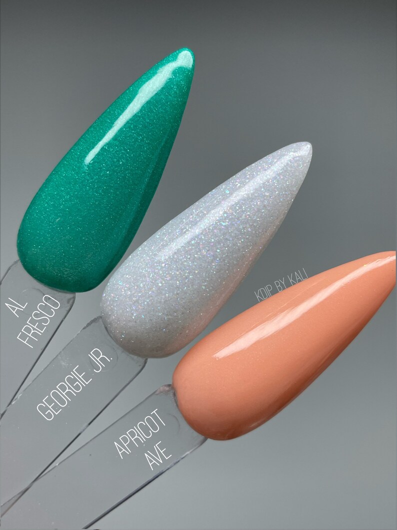 May include: Three nail polish colors in small bottles, from left to right: teal green, white with iridescent glitter, and peach. The bottles are labeled "Al Fresco", "Georgie Jr.", and "Apricot Ave".
