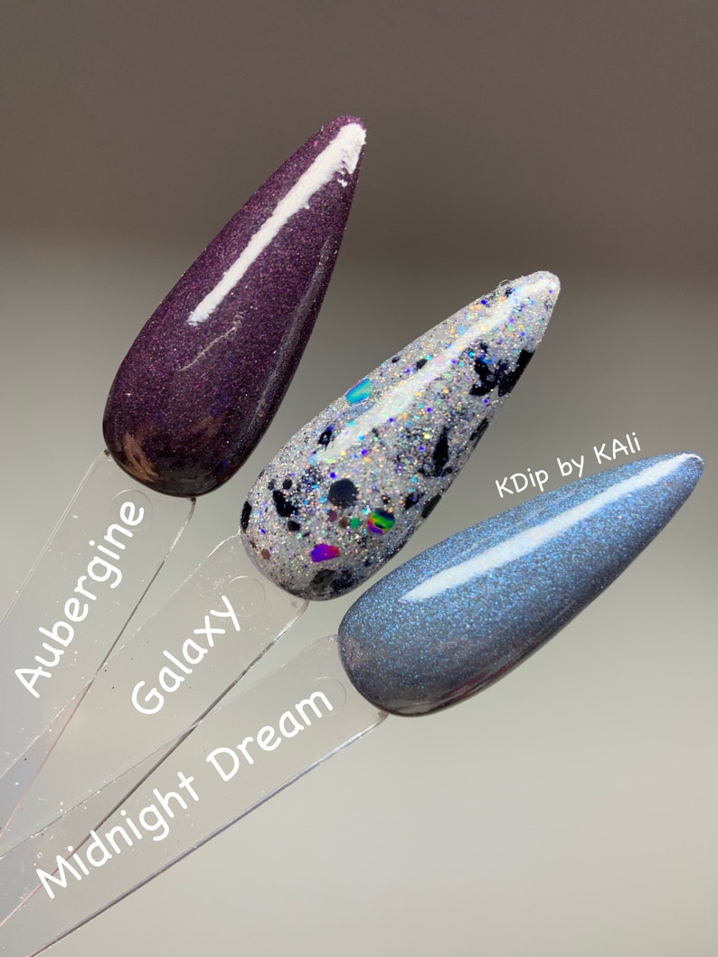 May include: Three nail polish swatches on clear sticks. The colors are Aubergine, Galaxy, and Midnight Dream. Aubergine is a dark purple with glitter. Galaxy is silver with black and iridescent glitter. Midnight Dream is a shimmery blue-gray.