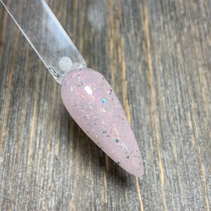 K&ouml;nnte beinhalten: Nahaufnahme eines helllilafarbenen Nagellacks mit schimmerndem Glitzer. Der Lack ist auf einen durchsichtigen Nagelstab aufgetragen.