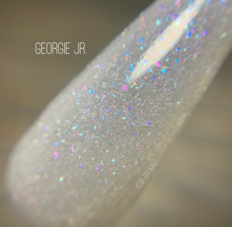 May include: A close-up of a clear nail polish bottle with a white iridescent glitter finish. The bottle is labeled "Georgie Jr." and "KOP BY KALI".