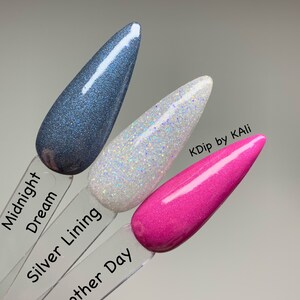 May include: Three nail polish swatches in Midnight Dream, Silver Lining, and Another Day. Midnight Dream is a dark blue with glitter. Silver Lining is white with iridescent glitter. Another Day is a bright pink. The brand name is KDip by KAli.