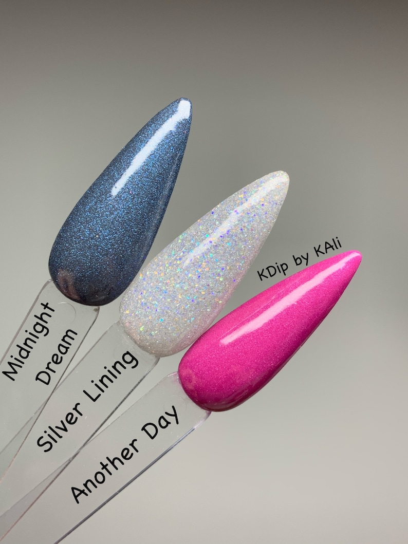 May include: Three nail polish swatches in Midnight Dream, Silver Lining, and Another Day. Midnight Dream is a dark blue with glitter. Silver Lining is white with iridescent glitter. Another Day is a bright pink. The brand name is KDip by KAli.