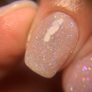 May include: Close-up of a light pink nail with a clear iridescent glitter top coat. The glitter is very fine and creates a shimmering effect. The nail is slightly curved and has a white design on top of the glitter.
