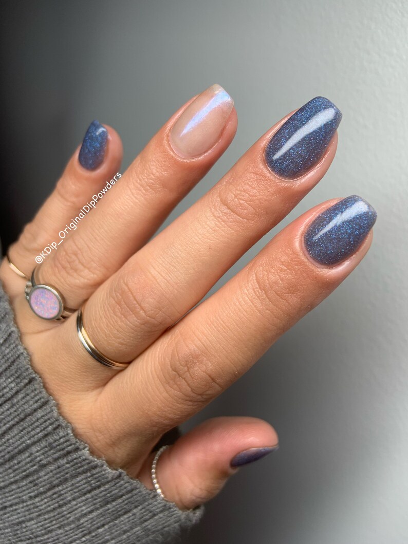 May include: Close-up of a hand with manicured nails. Three nails are painted a shimmering blue-grey, while one is a nude color with a subtle iridescent sheen. The hand is adorned with several rings, including a gold band and a ring with a pink stone.