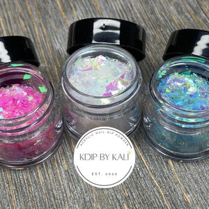 May include: Three small jars of iridescent glitter, each with a different color: pink, white, and blue. The jars are labeled "Acrylic Nail Dip Powder" and "Kdip by Kali, Est. 2020".