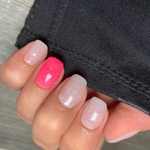 May include: Close-up of a hand with five fingernails painted with glitter nail polish. The nails are painted in a gradient of pink and white glitter. The hand is resting on a black fabric surface. The text "@KDip_OriginalDipPowders" is visible in the lower right corner of the image.