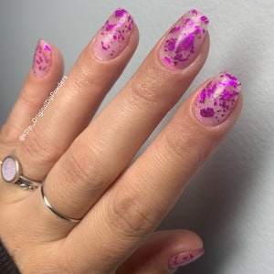 May include: Close-up of a hand with long, oval-shaped nails. The nails are painted with a clear base and adorned with vibrant pink foil flakes. The hand also features silver rings, including a ring with an opal stone.