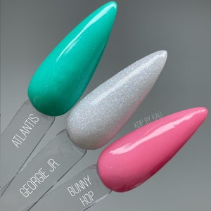 May include: Three nail polish colors on display. The colors are teal, white with glitter, and pink. The names of the colors are "Atlantis", "Georgie Jr.", and "Bunny Hop".