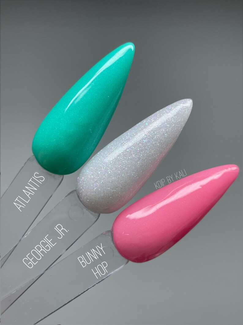 May include: Three nail polish colors on display. The colors are teal, white with glitter, and pink. The names of the colors are "Atlantis", "Georgie Jr.", and "Bunny Hop".