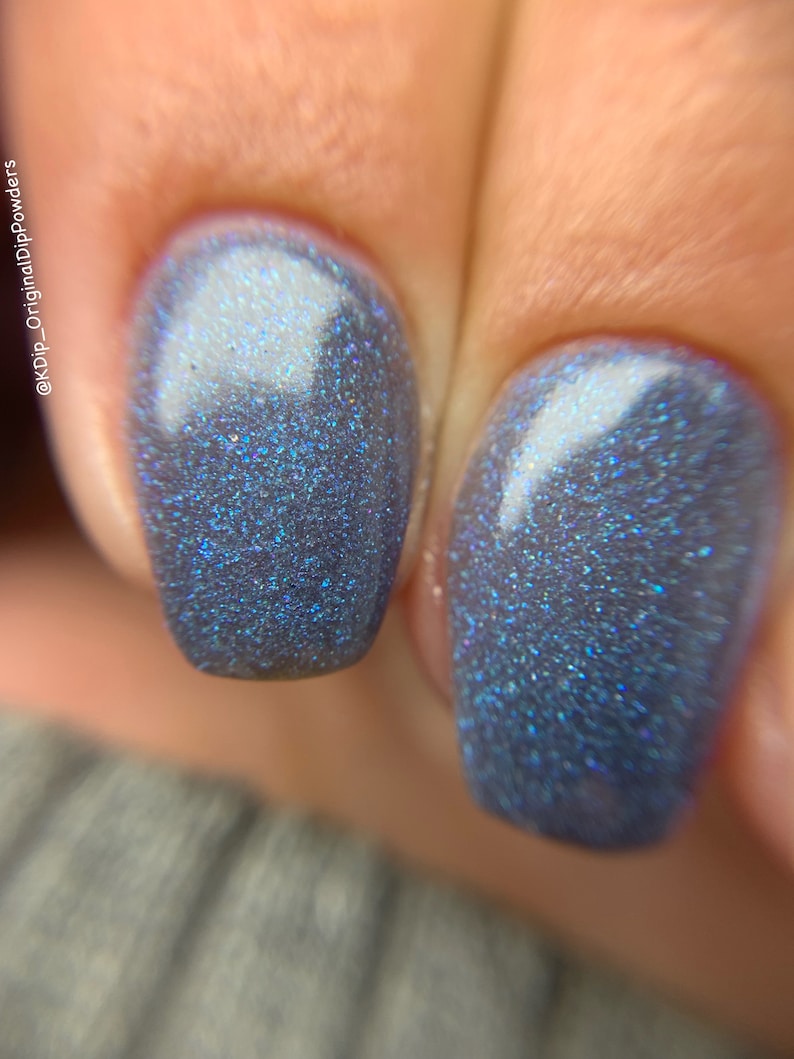 May include: Close-up of two fingernails painted with a shimmering, dark blue nail polish. The polish has a glittery, iridescent effect, reflecting shades of blue and purple. The nails are neatly shaped and well-manicured.