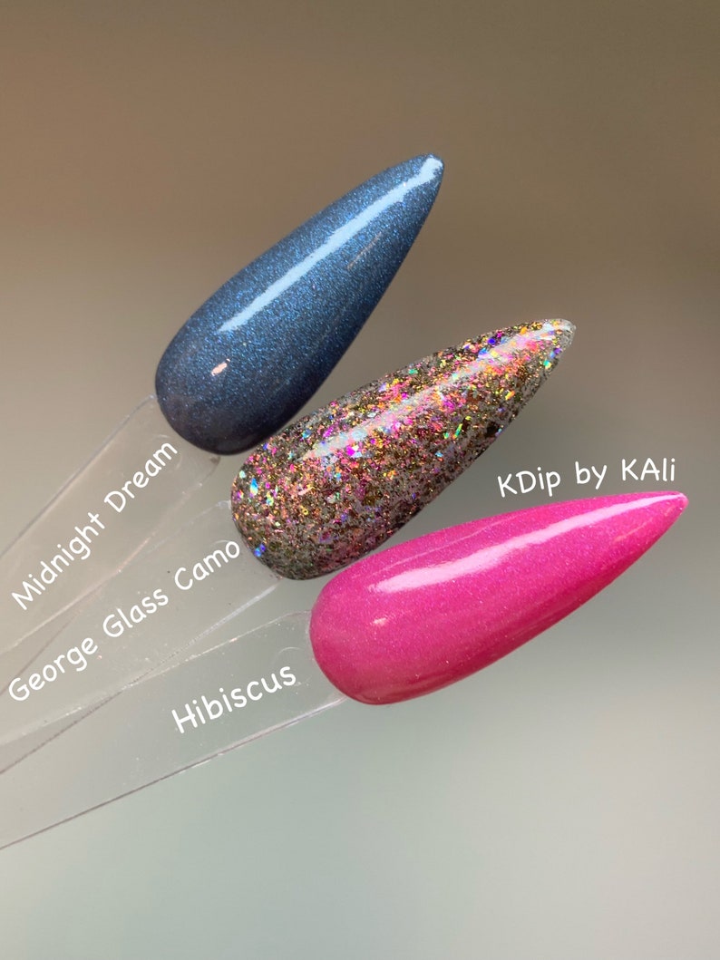 May include: Three nail polish swatches on clear sticks. The colors are Midnight Dream, a dark blue with shimmer; George Glass Camo, a glittery mix of gold, pink, and silver; and Hibiscus, a bright pink. The text "KDip by KALI" is visible.