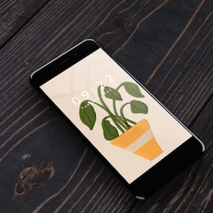 Könnte beinhalten: Ein schwarzes Smartphone zeigt eine digitale Illustration einer Topfpflanze mit grünen Blättern und einem gelben Topf. Der Bildschirm zeigt die Uhrzeit 09:22 und das Datum Dienstag, 19. Mai.