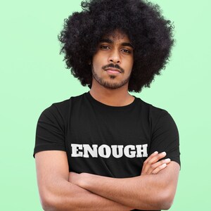 May include: A person wearing a black t-shirt with the word "ENOUGH" printed in white letters. The person is standing in front of a green background.