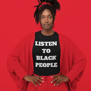 May include: A person wearing a black t-shirt with the text "LISTEN TO BLACK PEOPLE" in white letters. The person is wearing a red cardigan and red pants. The background is red.