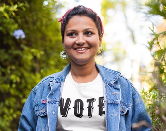 May include: A woman wearing a white t-shirt with the word "VOTE" printed in black, a denim jacket, and blue jeans. She is smiling and looking to the right.