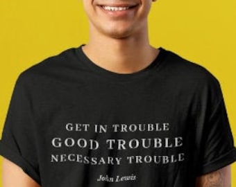 May include: A black t-shirt with white text that reads "Get in trouble, good trouble, necessary trouble - John Lewis". A person is wearing the t-shirt and smiling at the camera.