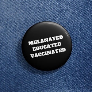 May include: A black button with white text that reads "Melanated Educated Vaccinated". The button is pinned to a blue denim jacket.