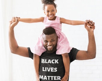 Puede incluir: Un hombre lleva una camiseta negra con el texto "Black Lives Matter" impreso en blanco. Lleva a una niña pequeña sobre sus hombros. La niña lleva un vestido rosa y sonríe.