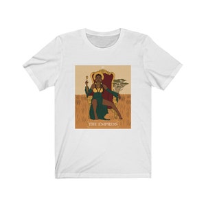 May include: A white t-shirt with a graphic of a tarot card depicting "The Empress". The card features a woman of color sitting on a throne with a crown and a scepter. The background is a field of tall grass.