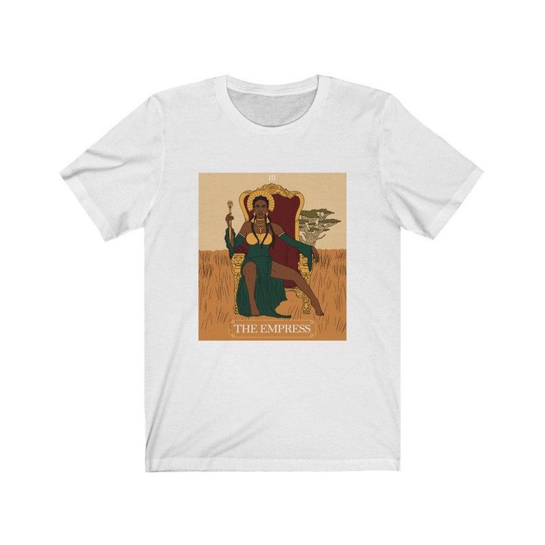 May include: A white t-shirt with a graphic of a tarot card depicting "The Empress". The card features a woman of color sitting on a throne with a crown and a scepter. The background is a field of tall grass.
