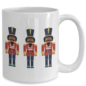 May include: White ceramic mug with a design of three black nutcracker soldiers wearing red and blue uniforms. The nutcrackers have black mustaches and beards.