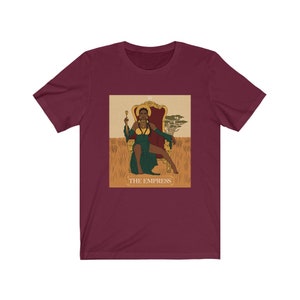 May include: A maroon t-shirt with a graphic of the Empress tarot card featuring a Black woman sitting on a throne. The card is surrounded by a gold frame and the text "THE EMPRESS" is printed below the image.