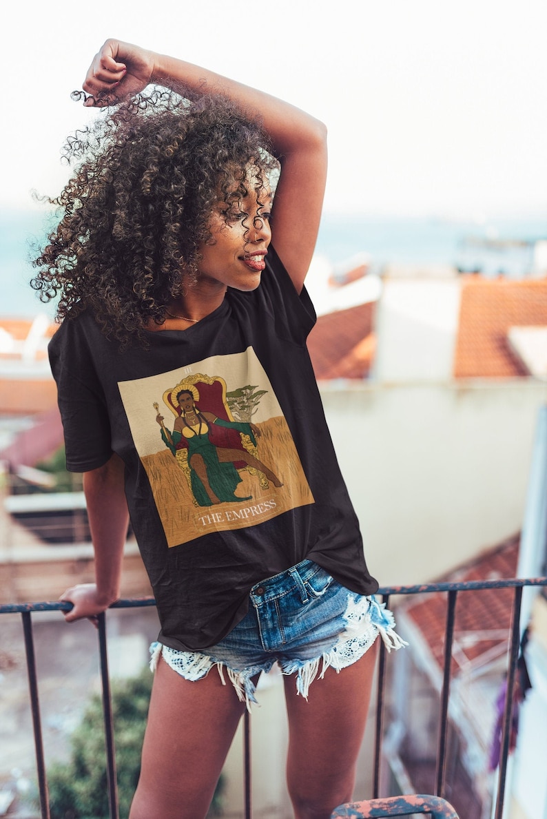 May include: A black t-shirt with a graphic of a woman sitting on a throne with the text "THE EMPRESS" underneath. The woman is wearing a green dress and a crown. She is holding a scepter in her right hand.