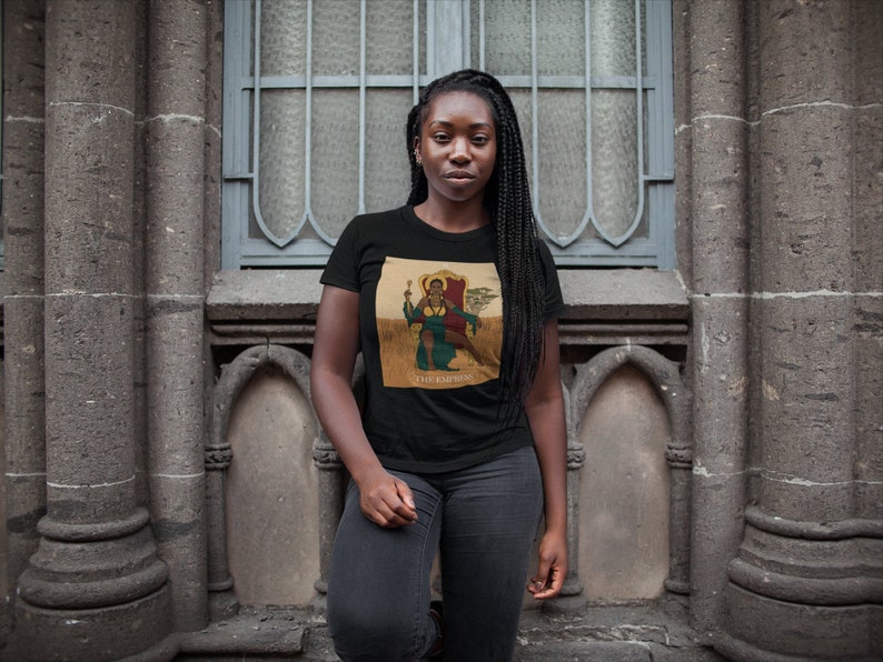 May include: A person wearing a black t-shirt with a graphic of a person sitting on a throne. The graphic is in a cartoon style and features a person with brown skin, a green shirt, and a gold crown. The text "The Empress" is below the graphic.