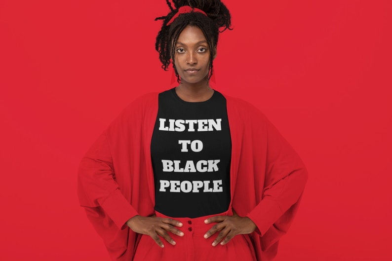 May include: A person wearing a black t-shirt with the text "LISTEN TO BLACK PEOPLE" in white letters. The person is wearing a red cardigan and red pants. The background is red.