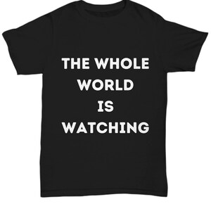Op de afbeelding: Zwart T-shirt met witte tekst die "THE WHOLE WORLD IS WATCHING" zegt.