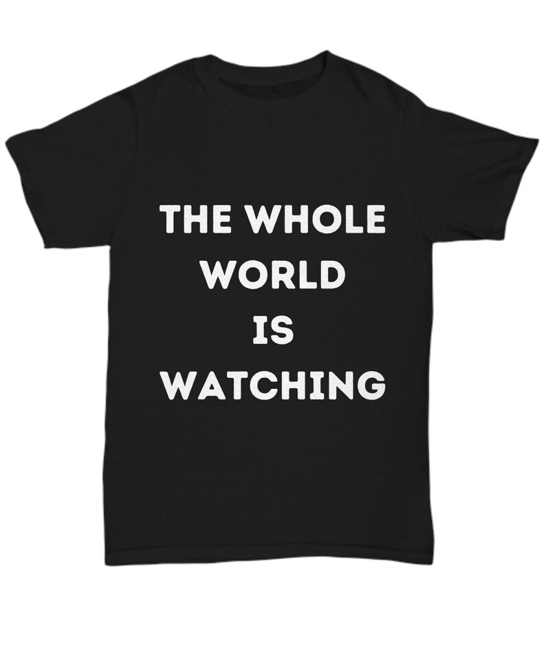 Op de afbeelding: Zwart T-shirt met witte tekst die "THE WHOLE WORLD IS WATCHING" zegt.