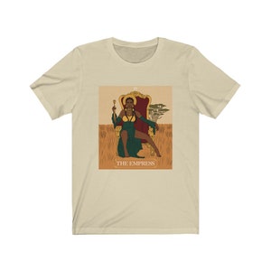 May include: A beige t-shirt with a graphic of the Empress tarot card featuring a Black woman sitting on a throne in a green dress. The card is surrounded by a gold frame and the text "THE EMPRESS" is printed below the image.