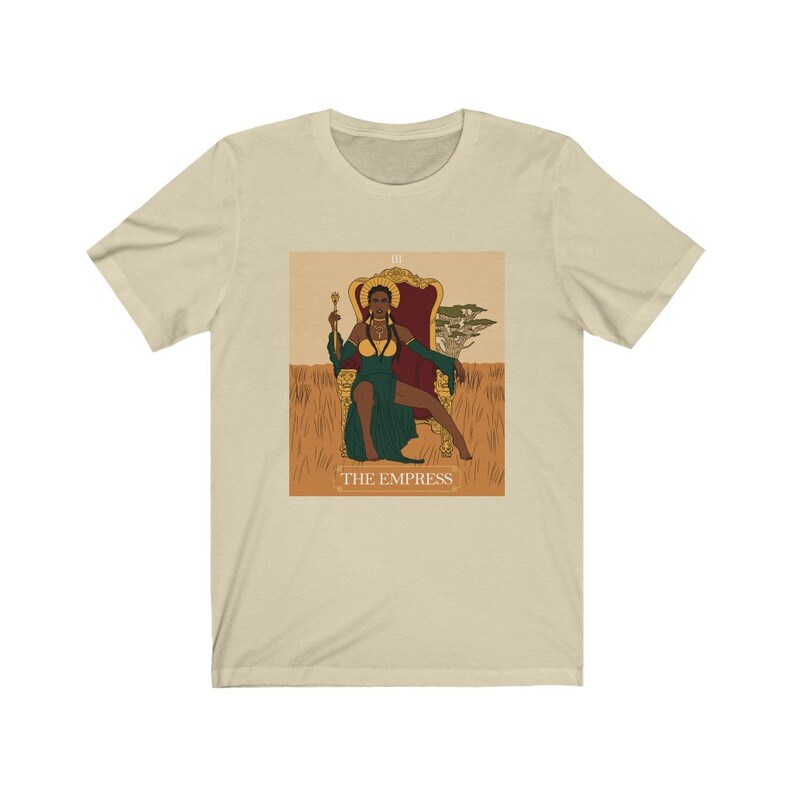 May include: A beige t-shirt with a graphic of the Empress tarot card featuring a Black woman sitting on a throne in a green dress. The card is surrounded by a gold frame and the text "THE EMPRESS" is printed below the image.
