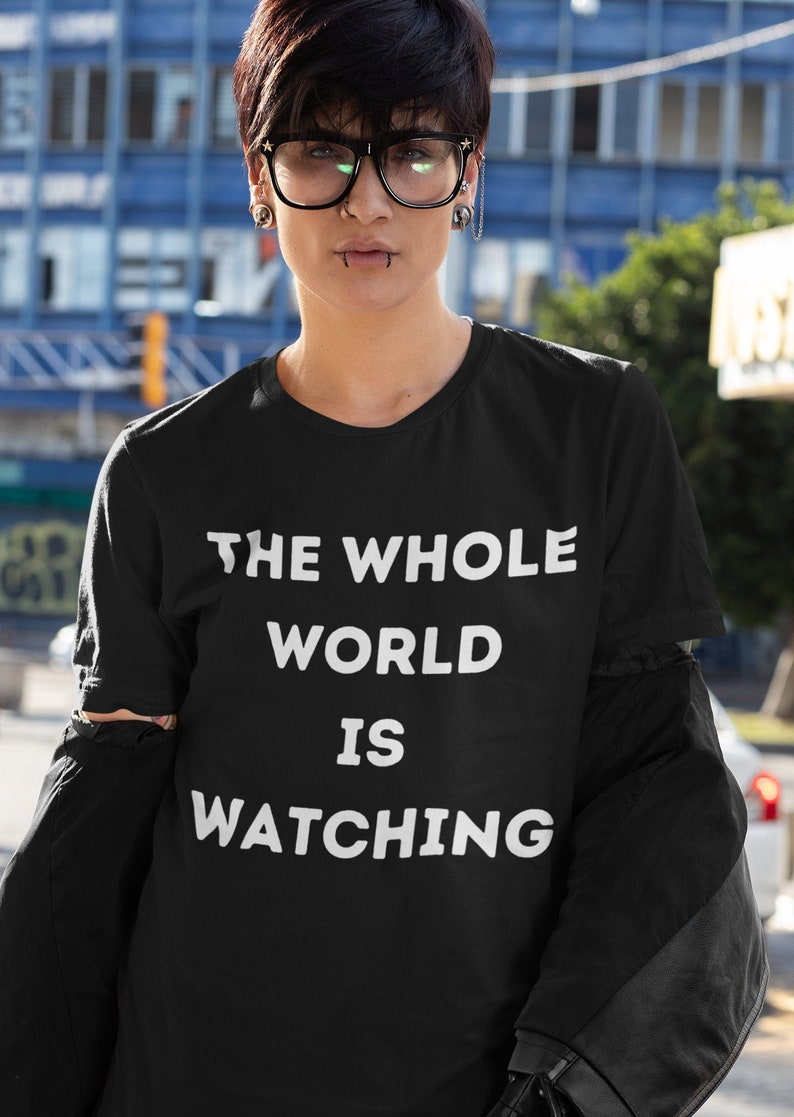Op de afbeelding: Een zwart T-shirt met witte tekst die "THE WHOLE WORLD IS WATCHING" zegt.